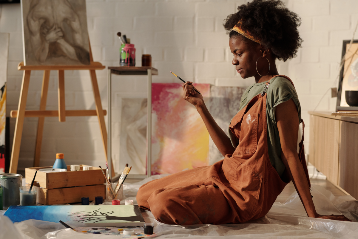Woman in overalls sits on art studio floor, holding a paintbrush while creating a painting.