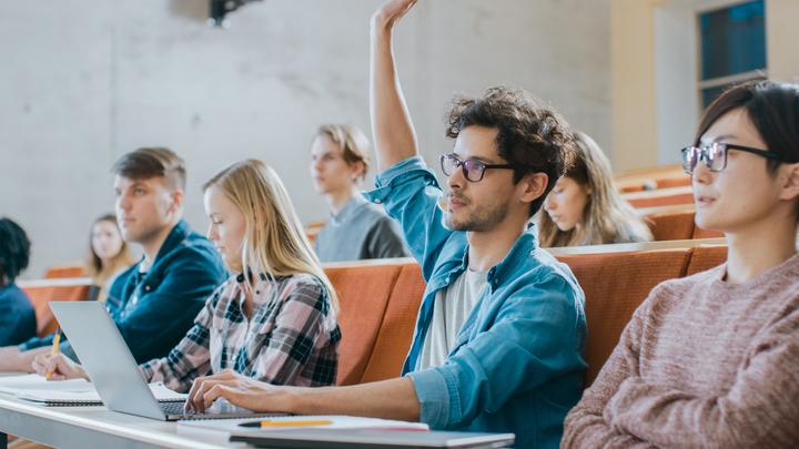 A young man raises his hand to ask a question during a university lecture with other students.