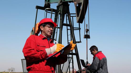 Petroleum engineer in red hard hat writes on clipboard near pump jack while colleague uses laptop.