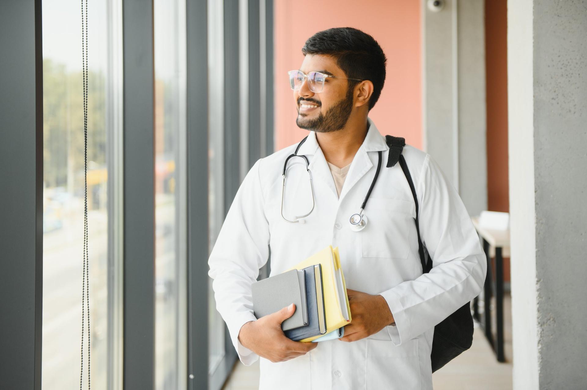 Smiling young medical student in lab coat and stethoscope stands by window holding textbooks.
