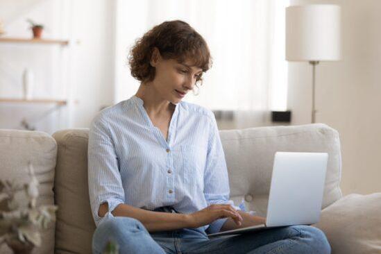Woman sits cross-legged on couch typing on laptop at home, representing writing a cover letter.