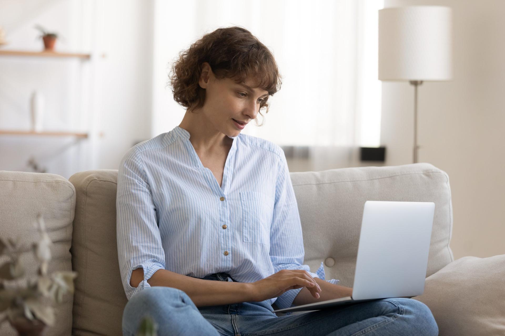 Woman sits cross-legged on couch typing on laptop at home, representing writing a cover letter.